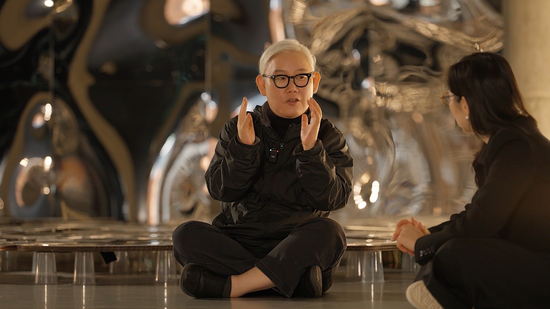 전시장 바닥에 앉아 손짓을 하며 작품에 대해 설명하고 있는 작가의 모습